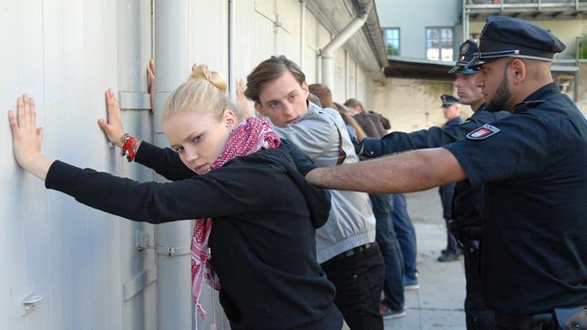 Eine junge Frau (Eva Nürnberg) und ein junger Mann (Alexander Finkenwirth) stehen mit anderen jungen Menschen mit den Händen an einer Wand. Hinter ihnen stehen Polizisten.