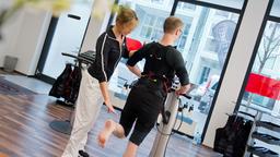 Eine Trainerin weist in einem Sportstudio einen Mann in Sportkleidung mit einer Weste, an der Kabel befestigt sind, vor einem Sportgert stehend, an.
