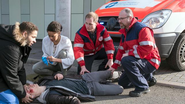Auf der Flucht vor Matteo (Mike Adler) wird Dennis (Elias Kaßner, liegend.) von einem Rettungswagen angefahren. Matteo und Vivi (Jane Chirwa, 2.v.r. mit Komparsen) kümmern sich sofort um den verletzten Jungen.
