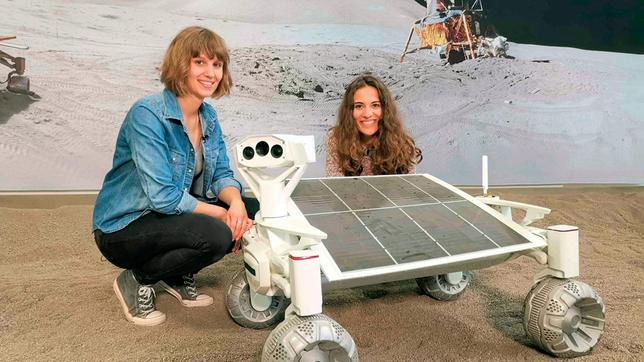 Mona (r) und die Wissenschaftlerin Nadine sitzen neben einem Rover, der eines Tages auf dem Mond umherfahren soll.