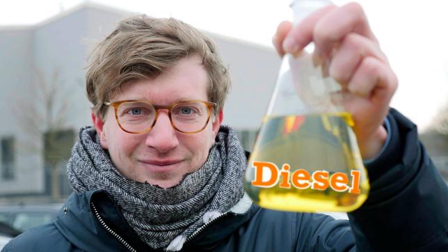 Robert hält einen Erlenmeyerkolben in der Hand. Darin ist eine gelbe Flüssigkeit. Auf dem Kolben steht der graphische Schriftzug „Diesel“.