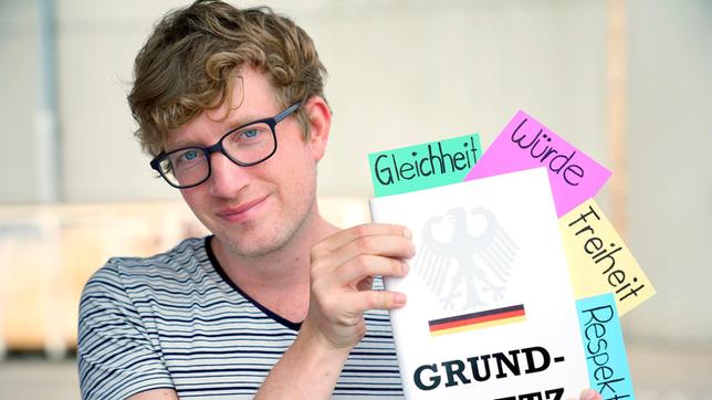 Robert mit einer Ausgabe des deutschen Grundgesetzes. Daraus hervor schauen bunte Karten mit den Grundrechten Gleichheit, Würde, Freiheit und Respekt.