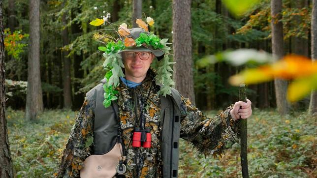 Bild: WDR Robert steht im Tarnoutfit im Wald. Bei sich trägt er Outdoor Ausrüstung wie Kompass, Fernglas und Wanderstock.