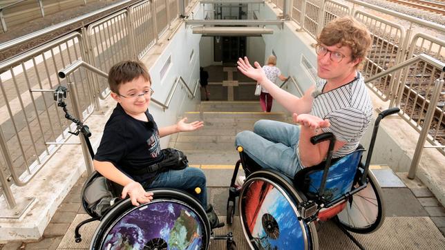 Bild: WDR/tvision Robert und Timon (l) stehen vor den Treppenstufen am Troisdorfer Bahnhof. Beide sitzen im Rollstuhl – Timon seit er 3 Jahre alt ist, Robert nur heute.