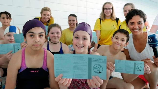 Bild: WDR/tvision Siham (r) gemeinsam mit den Schülern der Schwimm-AG des Leibniz-Gymnasiums Essen. Einige der Schüler halten ihren Jugend-Schwimmpass in die Kamera.