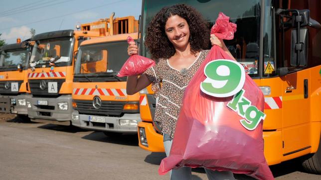 Siham mit einem kleinen und einem großen Müllsack auf dem Gelände der Stadtwerke Neuss. Im Hintergrund sind einige Müllfahrzeuge zu sehen.