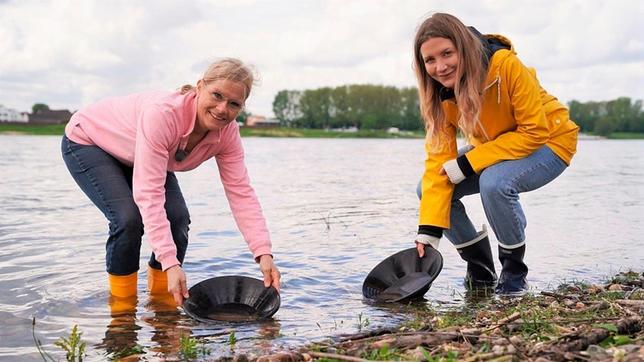 Um im Rhein Goldflitter finden zu können, braucht man einiges an Ausrüstung – zum Beispiel Goldwaschpfannen. – Jana und die Goldschmiedin Silke im Flussbett.