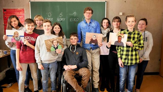 Robert mit einer Gruppe Schüler*innen vor einer Tafel. Sie halten einige großformatige Portraitfotos in den Händen.