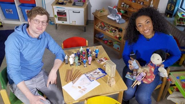 Robert sitzt mit Anne Chebu an einem Kindertisch, auf dem vielfältige und diverse Spielsachen liegen. Es gibt eine Schwarze und eine asiatische Puppe und Buntstifte in unterschiedlichen Hauttönen.