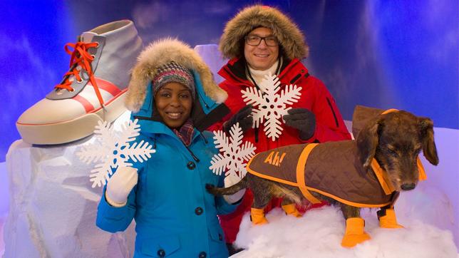 Shary und Ralph stehen mit einem Hund in einer Schnee-Kulisse. Sie tragen Anoraks, hinter ihnen liegt ein großer Schuh auf einem Eisblock.