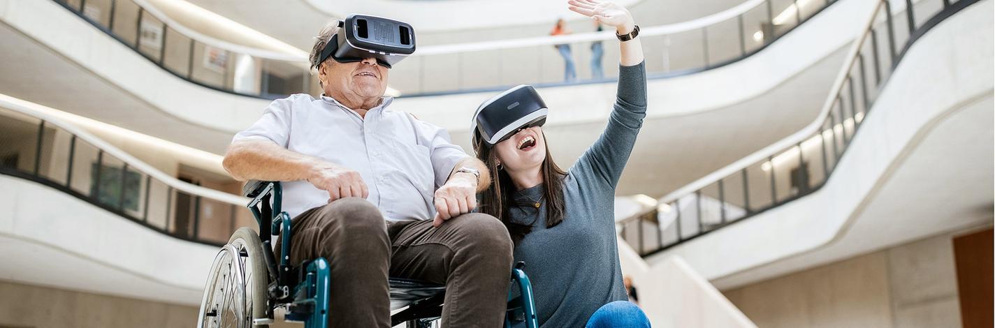 Freizeiterlebnis im virtuellen Raum: Ein älterer Mann im Rollstuhl und eine neben ihm knieende junge Frau mit Virtual-Reality-Brillen