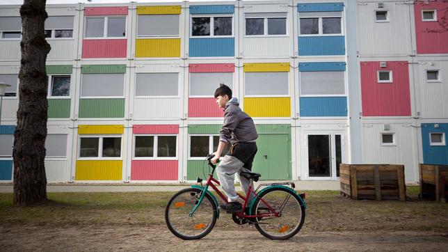 Kind fährt auf einem Rad vor aufeinandergestapelten Wohncontainern. 