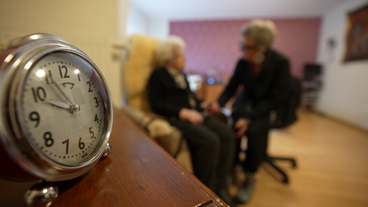 Im Vordergrund eine Uhr, im Hintergrund Pflegepersonal dass sich um eine Renterin kümmert.