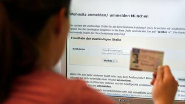 Bild: picture alliance / dpa | Paul Knecht Eine Frau sitzt vor dem Online-Verwaltungsportal einer Behörde und hält einen Personal- und einen Führerschein in ihrer rechten Hand.