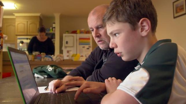 Vater und Sohn vor einem Laptop, Mutter in Hintergrund in der Küche.