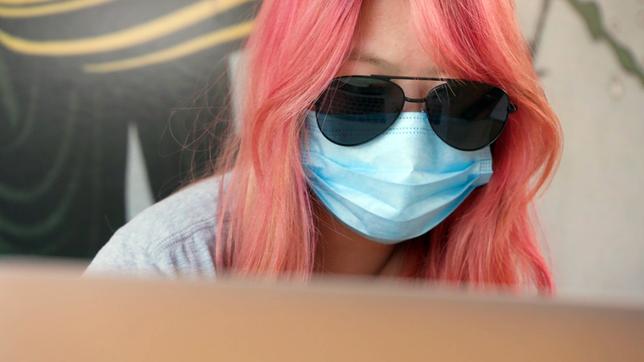 Eine Frau mit Sonnenbrille und Maske schaut auf den Bildschirm eines Laptops.