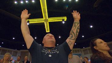 Ein Mann betet mit erhobenen Händen unter einem Kreuz.