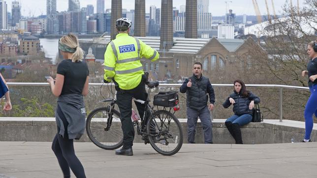 Ein Polizist auf einem Fahrrad hält an und spricht zwei sitzende Personen in einem Park in London an.