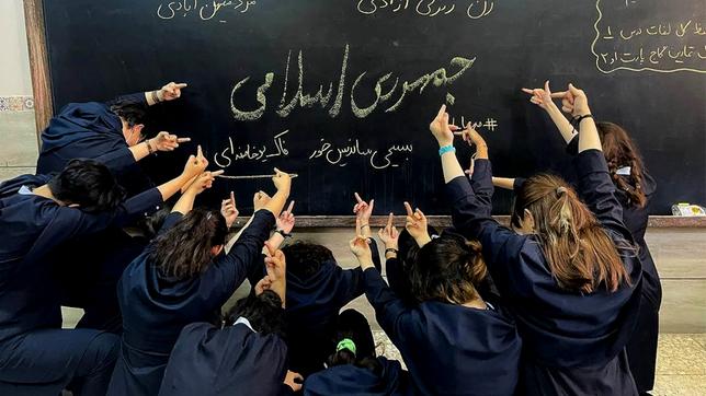 Schülerinnen mit offenen Haaren zeigen ihre Mittelfinger auf eine Schultafel mit einer Schrift.