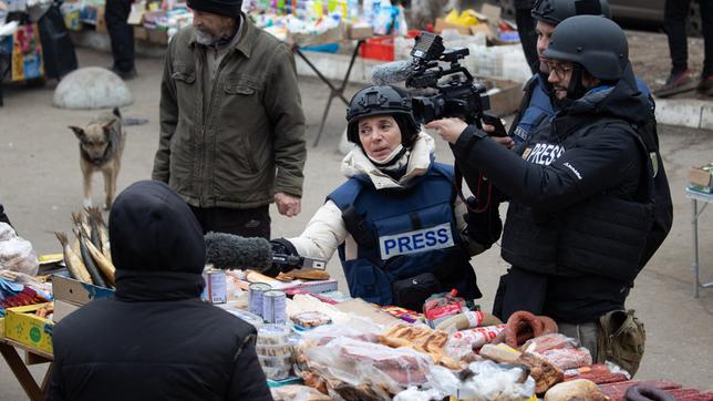 Drei Journalisten mit Helmen und Schutzwesten beim Dreh auf einem Markt