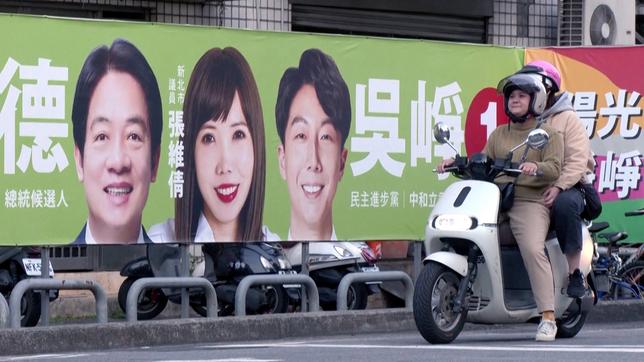 Ein Roller mit zwei Personen fährt an einem Wahlplakat vorbei.