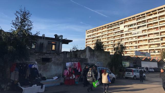 Menschen gehen eine Straße entlang. Im Hintergrund ein großes Wohnhaus im Sonnenschein.