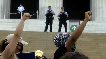 Zwei Frauen recken die Faust, im Hintergrund stehen Polizisten.