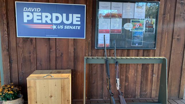 Wahlkampfschild an einer Holzwand, im Vordergrund zwei Waffen.
