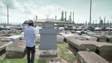 Mann steht auf Friedhof vor Grabstein, im Hintergrund Industrieanlagen