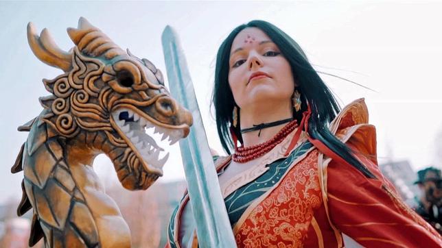 Eine junge Frau in einem fantastischen Kostüm und einem Schwert posiert neben dem Kopf eines Drachen.