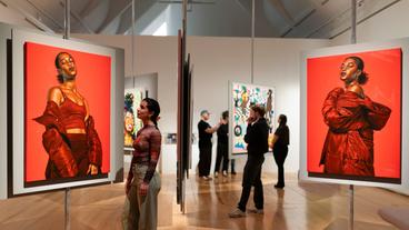 Personen in einem Ausstellungsraum betrachten Bilder, im Vordergrund eine Frau die auf orangefarbene Ölgemälde schwarzer Frauen schaut. 
