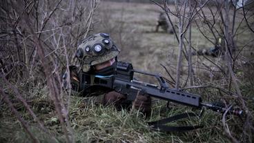 Ein Soldat liegt im Gebüsch und hält ein Gewehr im Anschlag.