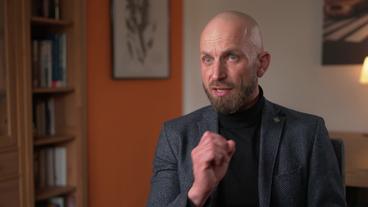 Ein mittelalter, Weißer Mann mit dunklem Bart und Glatze, in einem dunklen Jackett, sitzt auf einem Stuhl und gestikuliert mit geballter Faust.