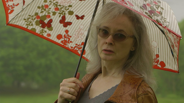 Bild: Screenshot Mary Gaitskill steht draußen mit eine Sonnebrille und Regeschrim und schaut in die Kamera.