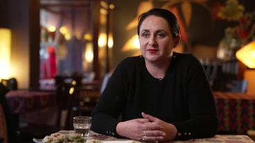 Bild: WDR Eine Frau mit zurückgekämmten schwarzen Haaren sitzt an einem Tisch.