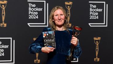 Bild: ddp/abaca press Eine Frau steht mit einem Buch und einer Trophäe vor einer Wand mit der Beschriftung "Booker Prize 2024"