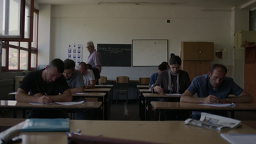 Bild: Screenshot Eine Gruppe an Menschen sitzen in einem Klassenzimmer und schreiben einen Test.