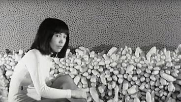 Bild: picture alliance / Everett Collection | Copyright Magnolia Pictures / Everett Collection Eine junge Yayoi Kusama sitzt vor einem ihrer Kunstwerke und schaut in die Kamera.