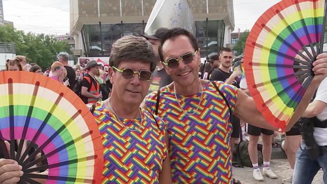 Zwei Männder in bunten T-Shirts und mit bunten Sonnenbrillen halten Fächer in Regenbogenfarben hoch.