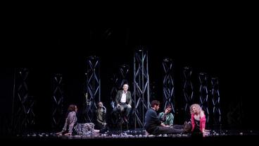 Eine dunkle Theaterbühne mit Aufbauten und sitzenden Schauspieler*innen