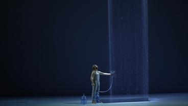 Ein Mann steht auf einer Theaterbühne in einem Lichtstrahl, der von der Decke fällt