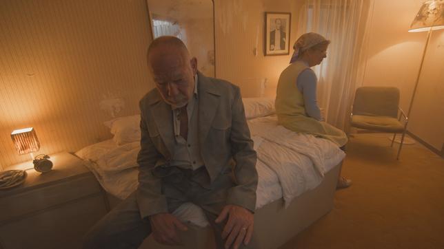 Ein Mann und eine Frau sitzen in einem düsteren Zimmer auf dem Bett