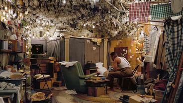 Mann sitzt in einem Raum, an der Decke hängen viele Lampen.