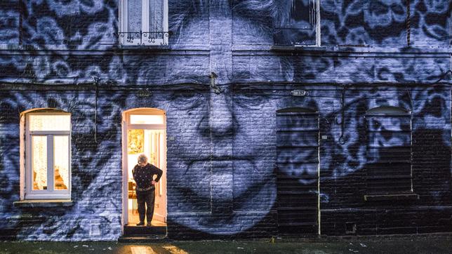 Bild: Agnès Varda-JR-Ciné-Tamaris, Social Animals 2016/Agnès Varda/JR eine Hauswand, auf dem übergroß das Foto einer Frau zu sehen ist, in der Haustür steht diese Frau