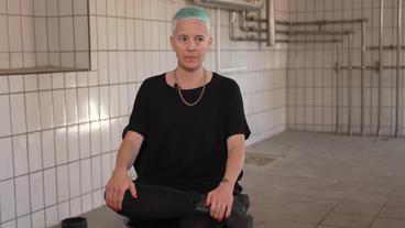 Bild: NDR Eine Frau mit kurzen, hellblau gefärbten Haaren sitzt in einem weiß gefließten Raum. Es ist die Künstlerin Candice Breitz.