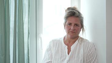 Eine mittelalte Frau mit blonden hochgesteckten Haaren, in weißer Bluse steht vor einem Fenster. Es ist die Autorin Gertraud Klemm.