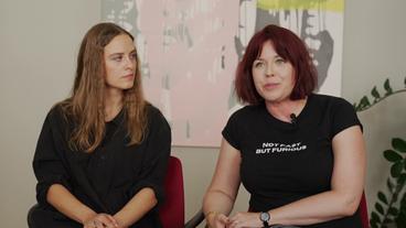 Eine junge Frau mit langen blonden Haaren und eine junge Frau mit halblangen dunkelroten Haaren sitzen nebeneinander. Es sind Eva Reisinger und Mareike Fallwickl.