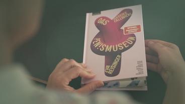 Zwei Hände halten das Buch "Das Pen!smuseum" und öffnen den Deckel. Das Cover zeigt eine geknotete Luftballonfigur, die an einen Penis erinnern soll.