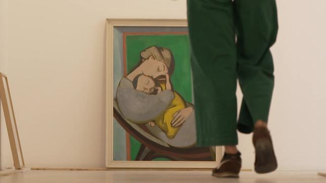 Ein Gemälde steht an eine Wand gelehnt, darauf eine Frau, die ein Kind hält, davor geht eine Person in grünen Hosen.