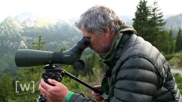 Ein Mann steht vor einer Alpenlandschaft und schaut in ein Fernglas.